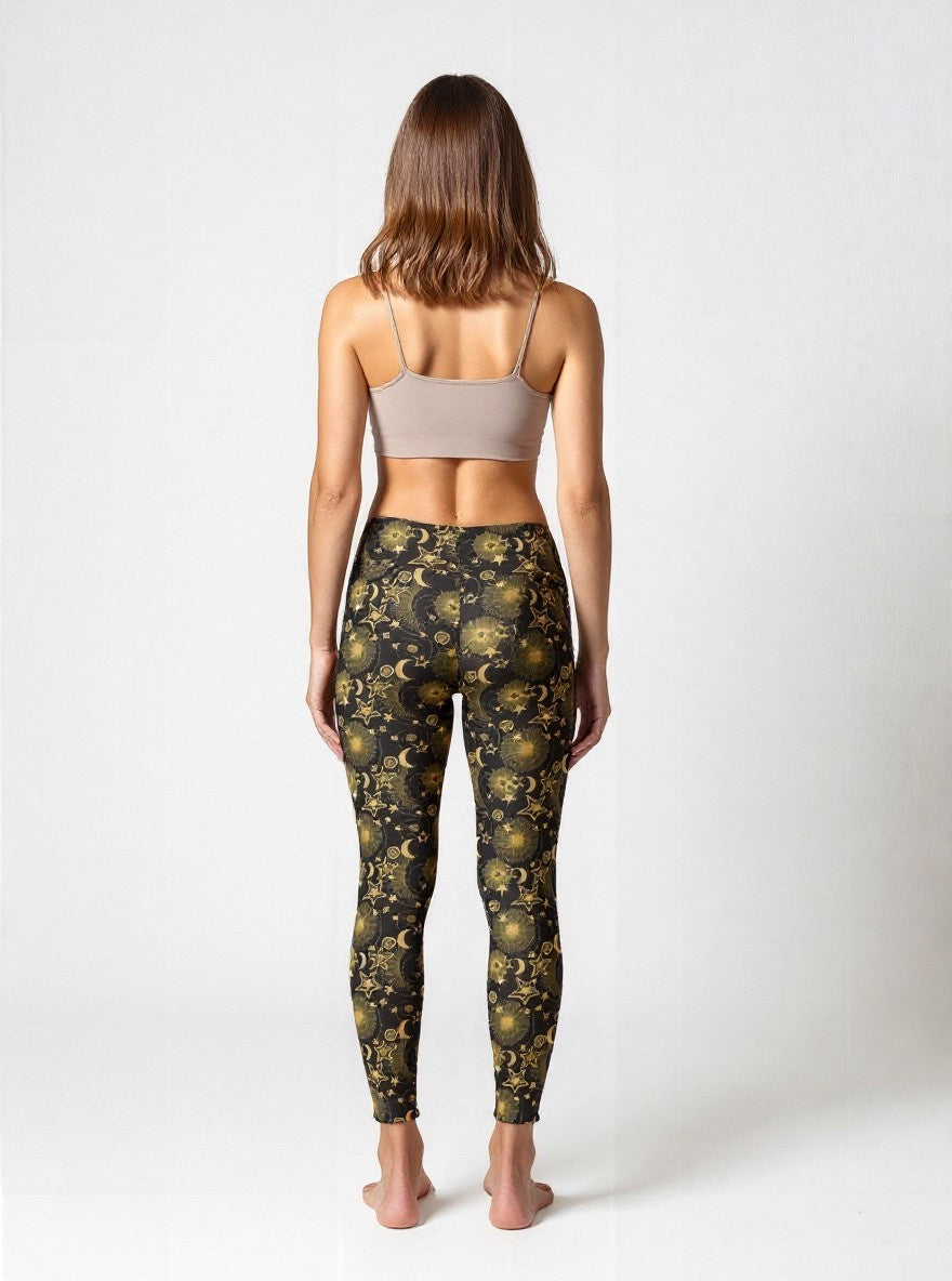 Woman wearing Midnight Halo yoga leggings with a celestial pattern featuring gold stars, suns, and moons on a black background, paired with a beige sports bra, standing barefoot against a plain white backdrop.