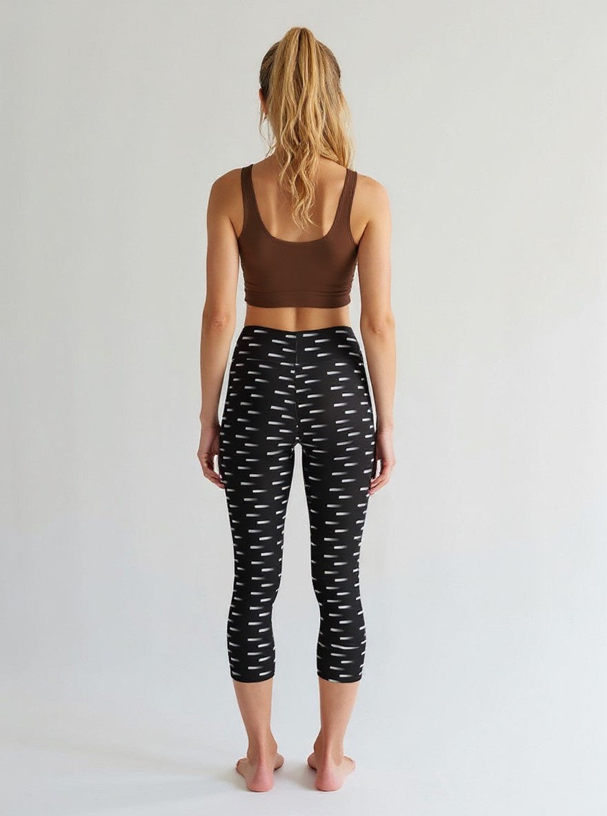 A woman is shown from the back wearing Nightfall Edit Capris, which feature a high-waisted, cropped fit with a black base and white dash print. She pairs the capri leggings with a brown sports bra, standing barefoot against a plain, light background.