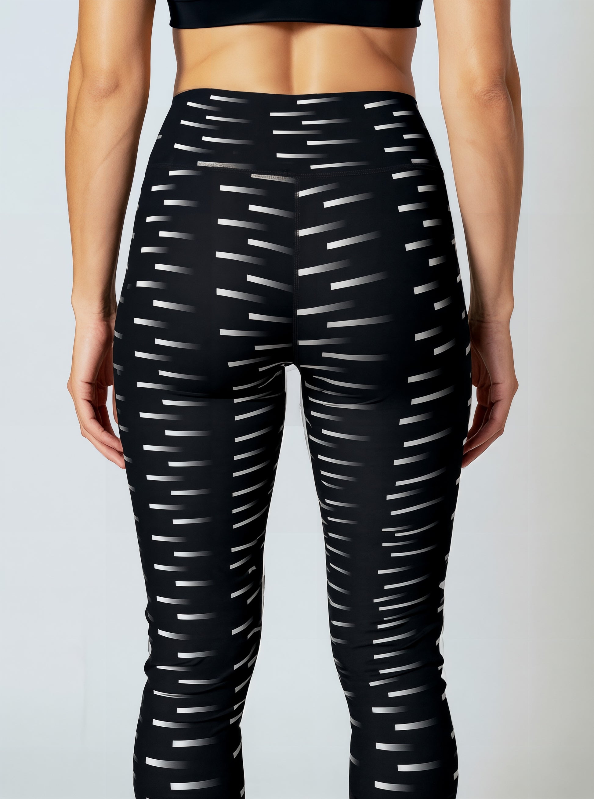 A back, close-up of a woman wearing Nightfall Edit leggings, showcasing the high-waisted fit and all-over black fabric with a white dash pattern. The image highlights the sleek waistband and the modern geometric print.