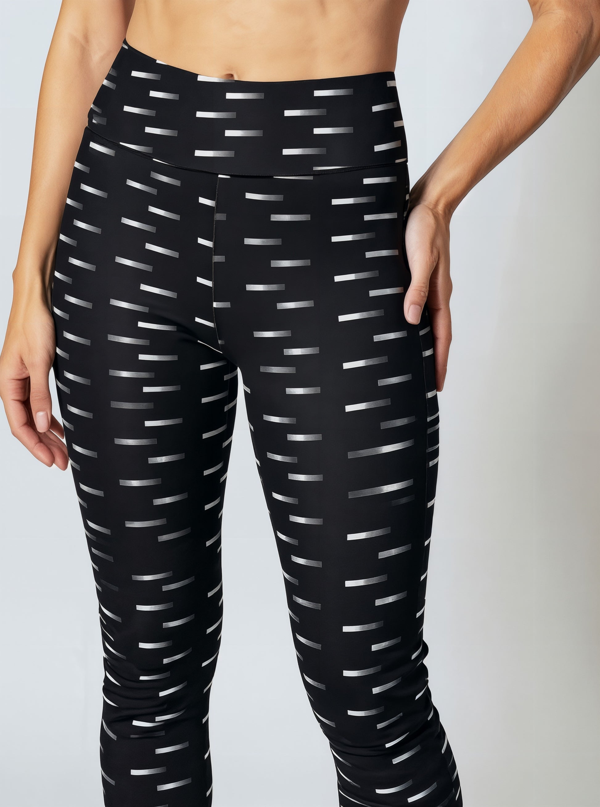 A close-up of a woman wearing Nightfall Edit leggings, showcasing the high-waisted fit and all-over black fabric with a white dash pattern. The image highlights the sleek waistband and the modern geometric print.