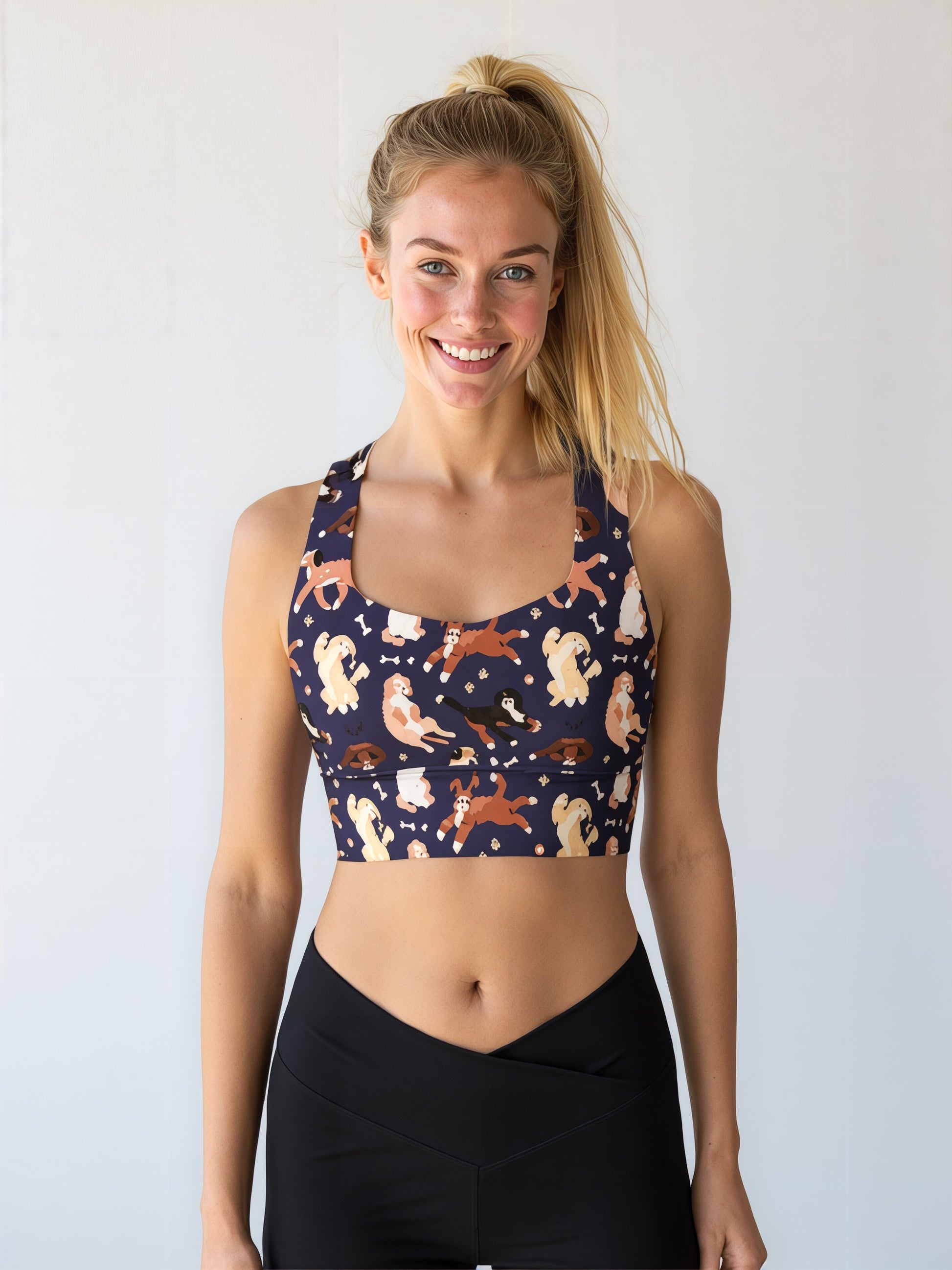 A woman smiles while wearing the Puppy Parade longline bra, featuring a playful print of various cartoon puppies in shades of cream, brown, and tan on a dark navy background. She pairs the bra with high-waisted black leggings and stands against a plain, light background.