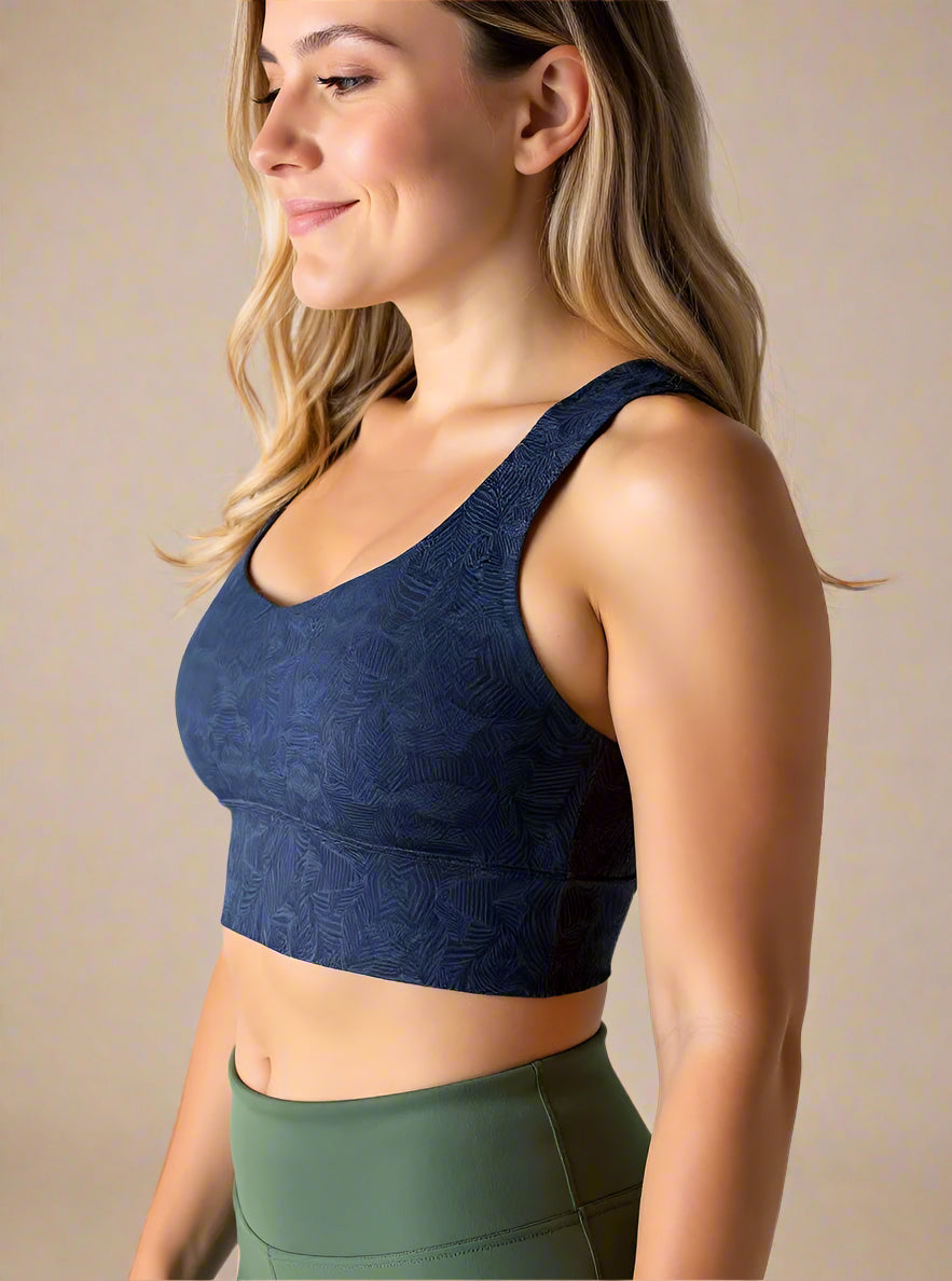 Side view of a woman with long, wavy blonde hair smiling and wearing a dark blue Shadowleaf longline sports bra with a subtle leaf pattern, paired with high-waisted olive green leggings. The image highlights the fit and texture of the sports bra.