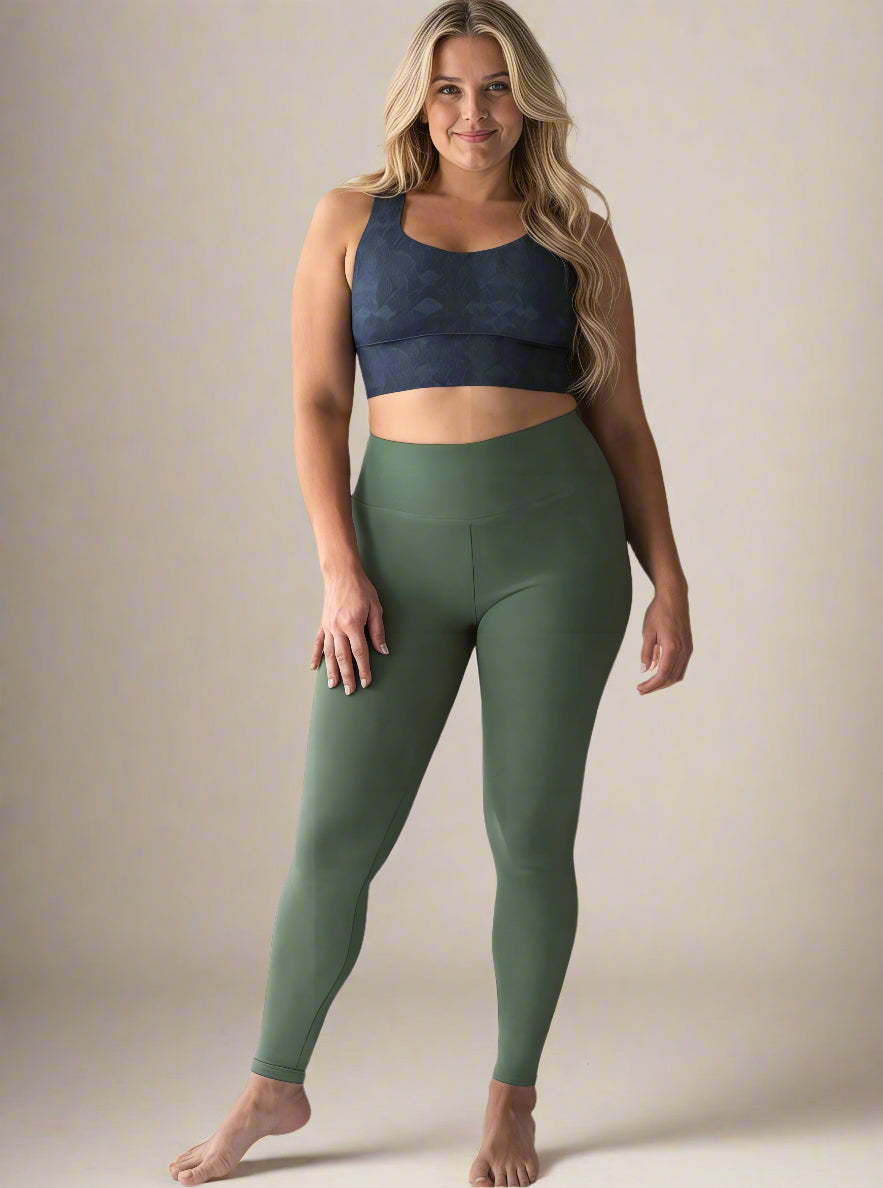 Curvy woman with long, wavy blonde hair wearing a dark blue Shadowleaf longline sports bra and high-waisted olive green leggings, standing barefoot and smiling confidently against a light background.