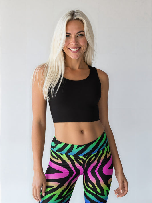 Smiling athletic woman with long straight white hair, fair but tanned skin, wearing a black crop top and vibrant technicolour zebra-print yoga leggings, standing against a plain light background.