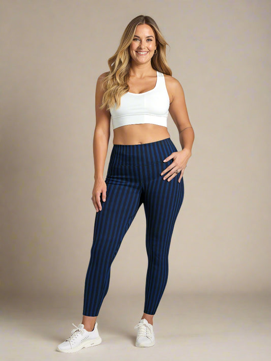 A woman with long, wavy blonde hair is smiling and standing against a plain background, wearing a white sports bra, blue and black vertically striped Wednesday pocket leggings, and white sneakers. One hand rests on her hip, showing the fit and style of the leggings.