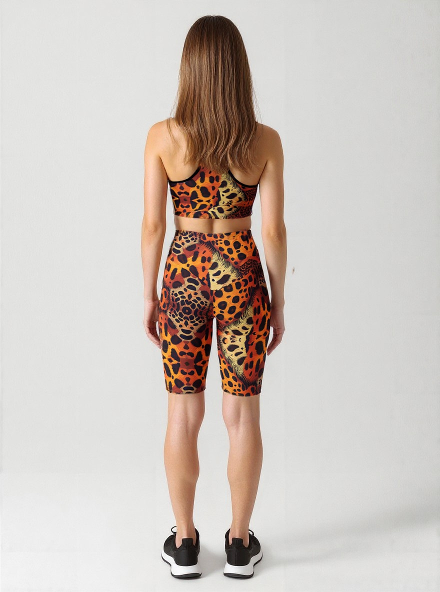 A woman is shown from behind, wearing a matching sports bra and Wildfire biker shorts set. The biker shorts feature a bold, all-over animal print pattern in orange, yellow, and black, with a high-waisted, fitted design that falls just above the knee. She is also wearing black athletic shoes, and the plain background highlights the vibrant, eye-catching details of the activewear.