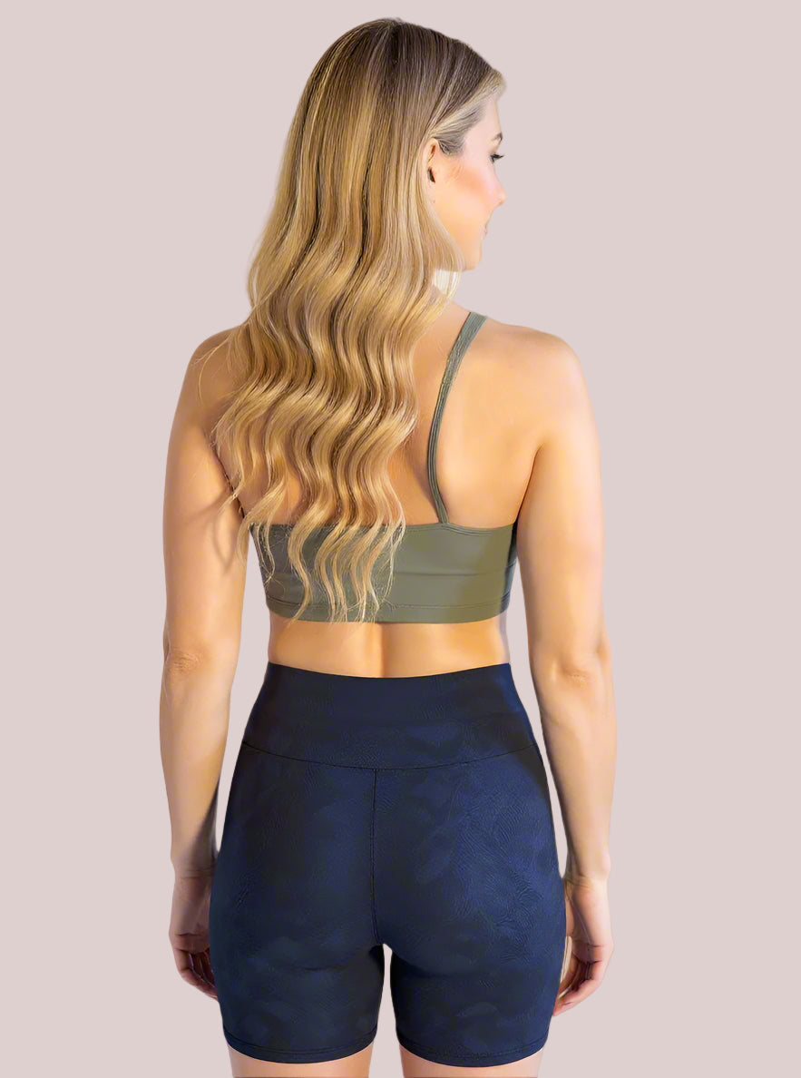 A woman with long, wavy blonde hair is shown from the back, wearing an olive green sports bra and high-waisted, dark navy Shadowleaf Yoga shorts with subtle textured detailing, standing against a light background.