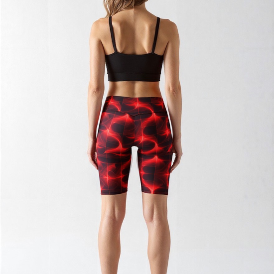 Back view of a woman wearing Blaze Ignite Biker Shorts with a bold red and black abstract pattern, paired with a black sports bra and white sneakers, standing against a plain background.