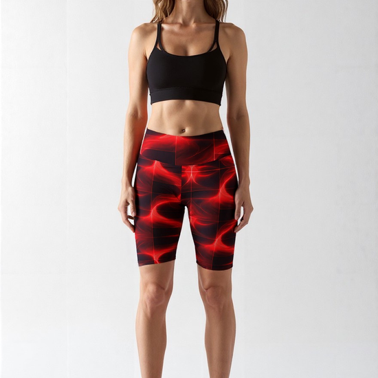 Front view of a woman wearing Blaze Ignite Bikers, featuring a vibrant red and black abstract pattern, paired with a black sports bra and white sneakers, standing against a plain light background.