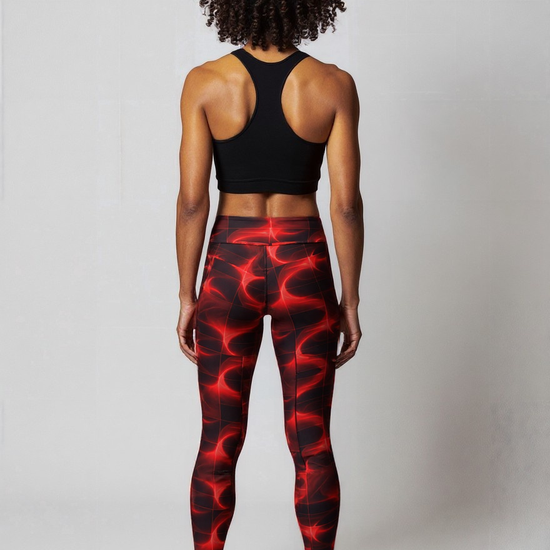 Back view of woman smiles confidently while wearing Blaze Ignite Yoga leggings, which showcase a striking red and black abstract pattern. She pairs the high-waisted, fitted leggings with a black athletic sports bra, standing in a relaxed pose against a plain light background.