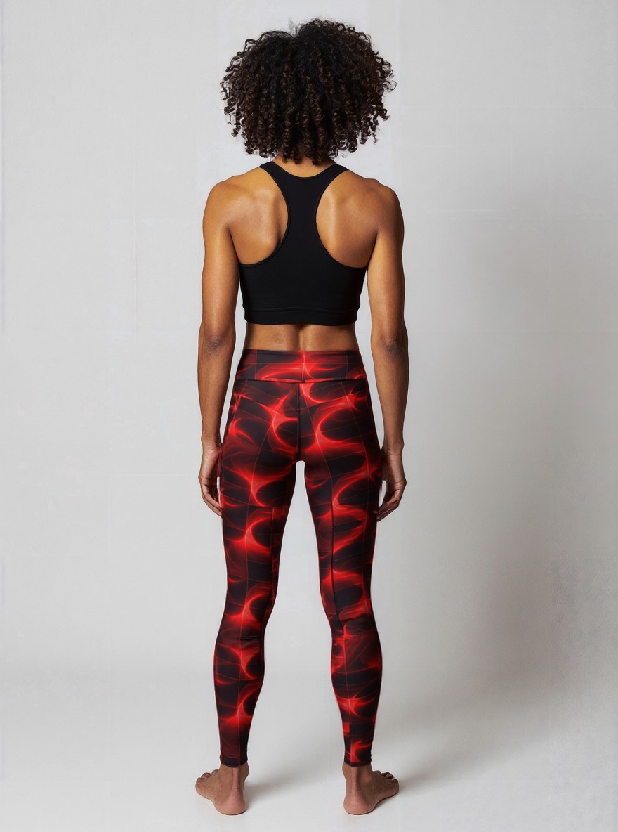 Back view of woman smiles confidently while wearing Blaze Ignite Yoga leggings, which showcase a striking red and black abstract pattern. She pairs the high-waisted, fitted leggings with a black athletic sports bra, standing in a relaxed pose against a plain light background.