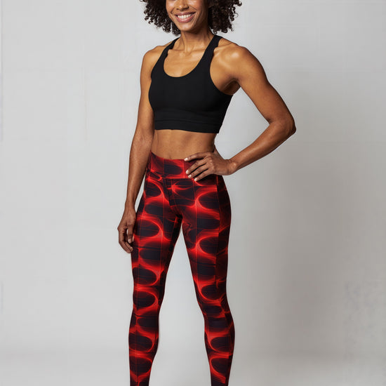 Full view of woman smiles confidently while wearing Blaze Ignite Yoga leggings, which showcase a striking red and black abstract pattern. She pairs the high-waisted, fitted leggings with a black athletic sports bra, standing in a relaxed pose against a plain light background.
