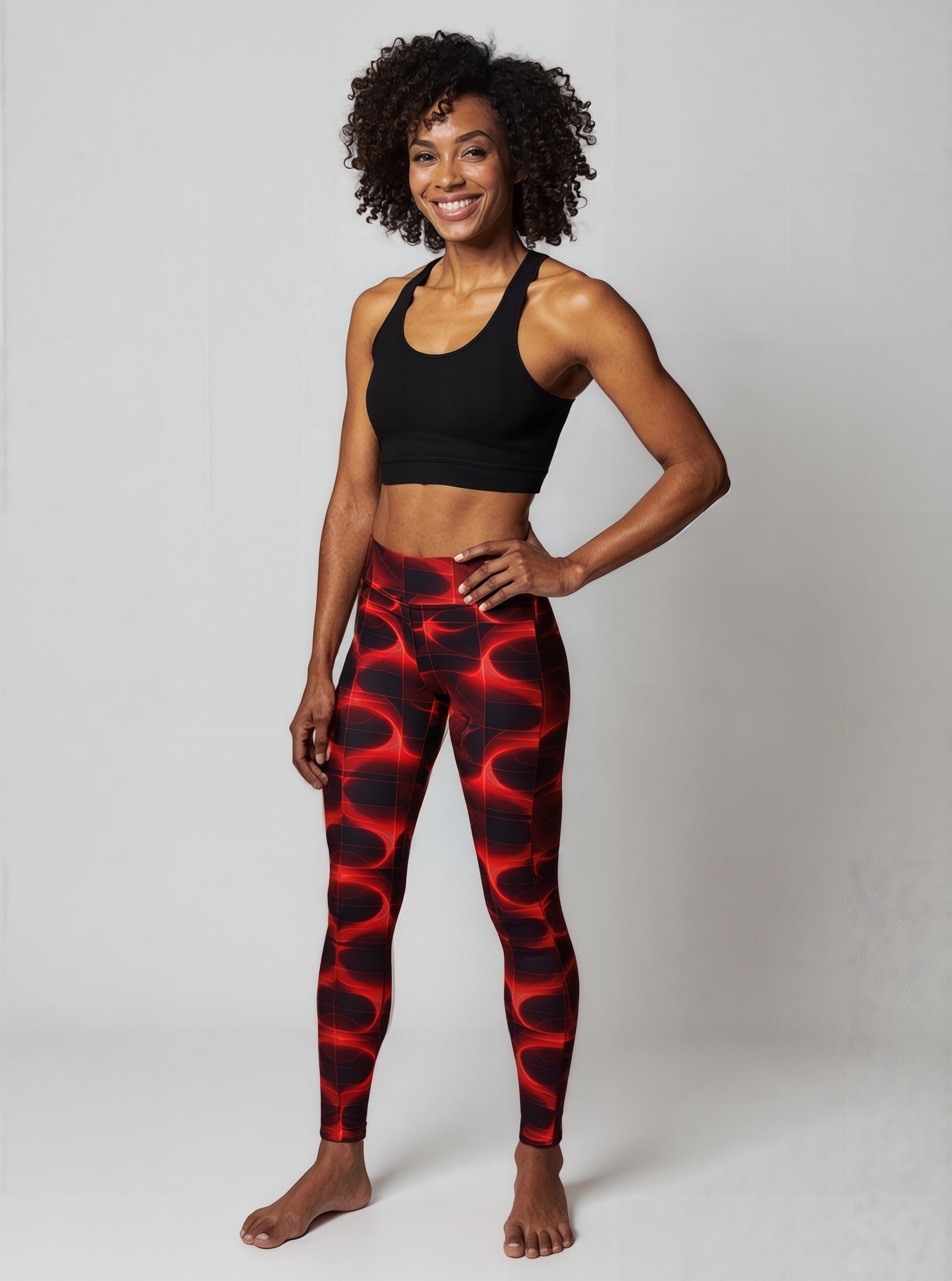 Full view of woman smiles confidently while wearing Blaze Ignite Yoga leggings, which showcase a striking red and black abstract pattern. She pairs the high-waisted, fitted leggings with a black athletic sports bra, standing in a relaxed pose against a plain light background.