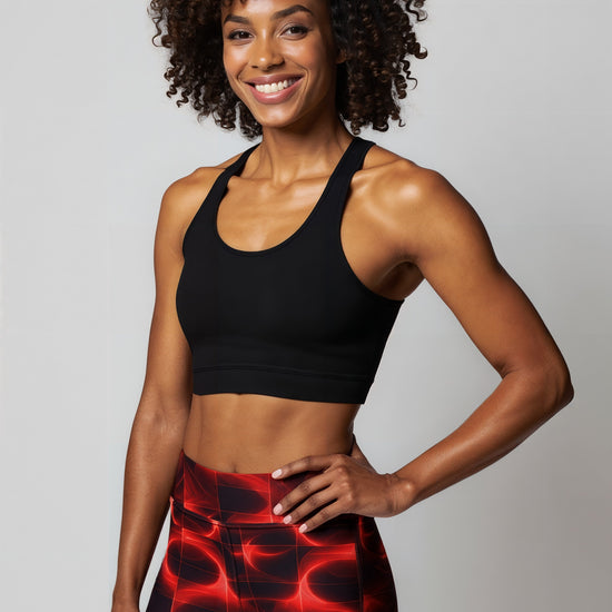 A woman smiles confidently while wearing Blaze Ignite Yoga leggings, which showcase a striking red and black abstract pattern. She pairs the high-waisted, fitted leggings with a black athletic sports bra, standing in a relaxed pose against a plain light background.