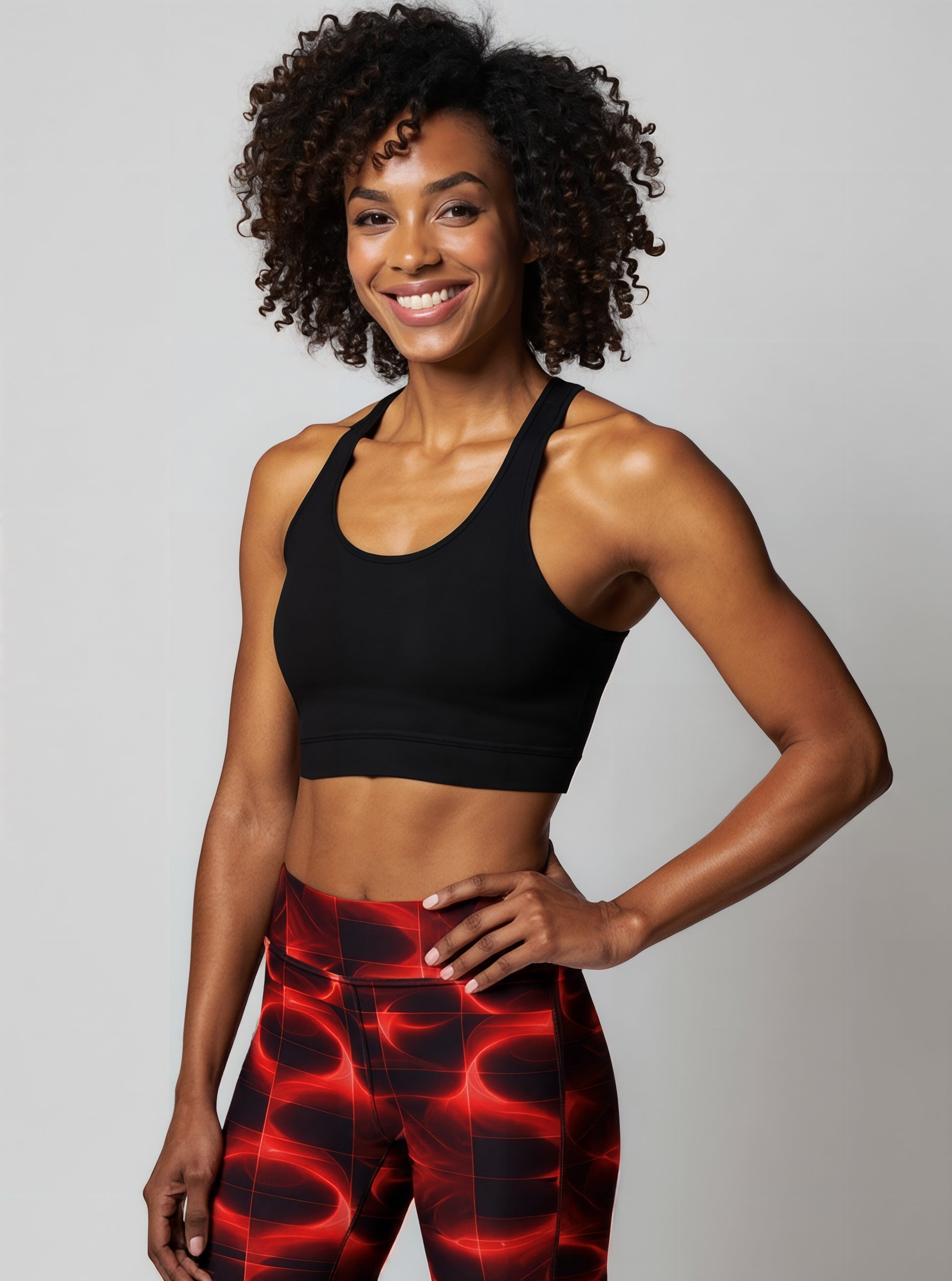 A woman smiles confidently while wearing Blaze Ignite Yoga leggings, which showcase a striking red and black abstract pattern. She pairs the high-waisted, fitted leggings with a black athletic sports bra, standing in a relaxed pose against a plain light background.