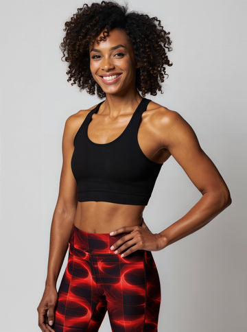 A woman smiles confidently while wearing Blaze Ignite Yoga leggings, which showcase a striking red and black abstract pattern. She pairs the high-waisted, fitted leggings with a black athletic sports bra, standing in a relaxed pose against a plain light background.