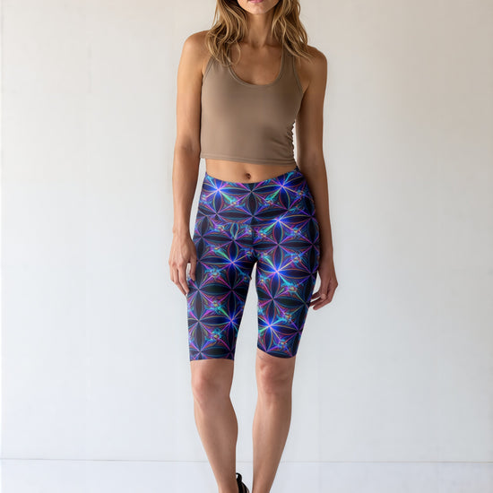 Woman wearing Blue Volt biker shorts with an electric blue and purple geometric print, paired with a tan crop tank and black athletic shoes, standing against a plain light background.