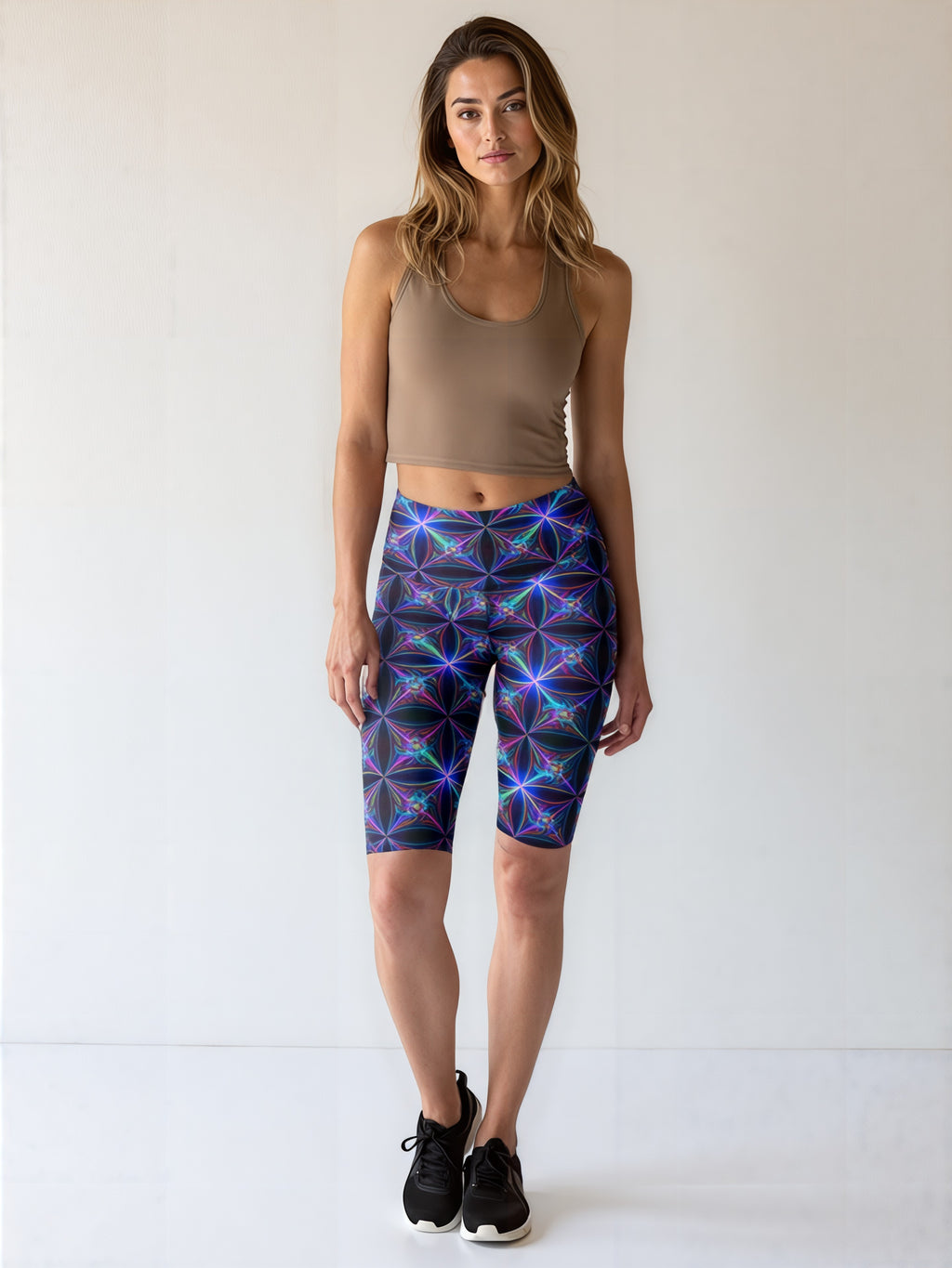 Woman wearing Blue Volt biker shorts with an electric blue and purple geometric print, paired with a tan crop tank and black athletic shoes, standing against a plain light background.
