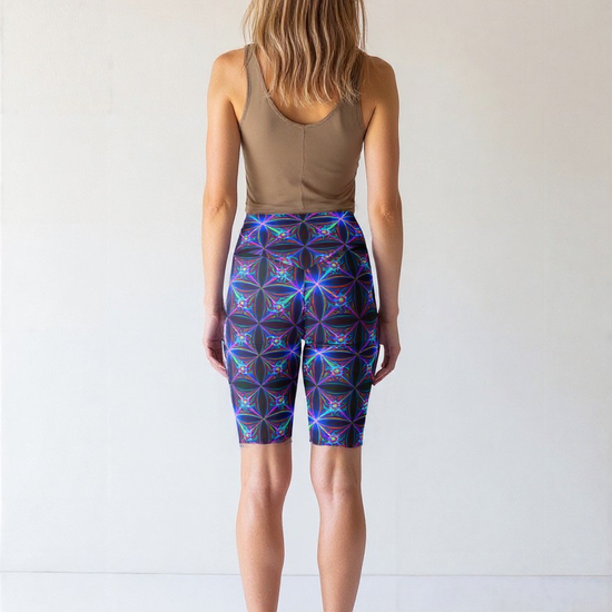 Back view of a woman wearing Blue Volt biker shorts with a vibrant electric blue and purple geometric pattern, paired with a taupe crop tank and black sneakers, standing against a plain light background.