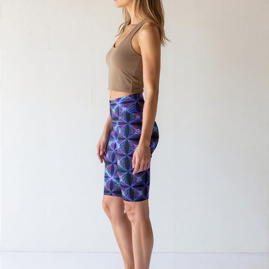 Side view of a woman wearing Blue Volt biker shorts featuring a vivid blue and purple geometric print, paired with a taupe crop top and black sneakers, standing against a plain light background.