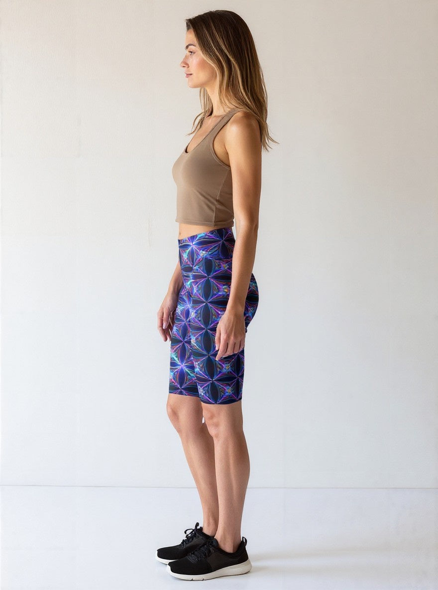 Side view of a woman wearing Blue Volt biker shorts featuring a vivid blue and purple geometric print, paired with a taupe crop top and black sneakers, standing against a plain light background.