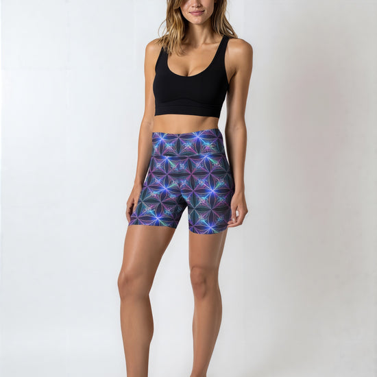 Woman wearing Blue Voltage Yoga shorts with an electric blue and purple geometric print, paired with a black sports bra and black sneakers, standing against a plain light background.