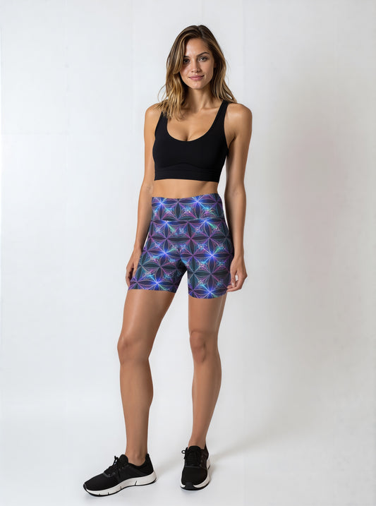 Woman wearing Blue Voltage Yoga shorts with an electric blue and purple geometric print, paired with a black sports bra and black sneakers, standing against a plain light background.