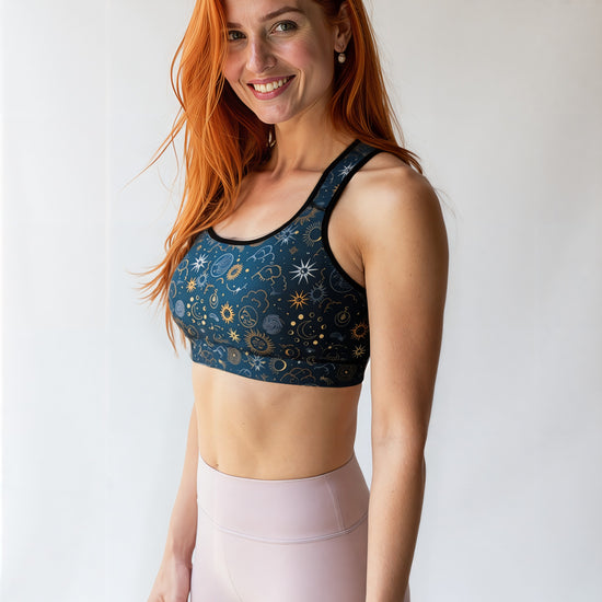 Smiling woman wearing the Celestial Essential Bra with a dark blue, star and moon pattern, paired with light pink high-waisted leggings, standing against a plain, light background.