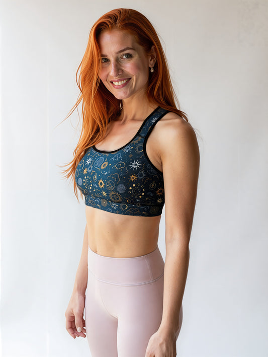 Smiling woman wearing the Celestial Essential Bra with a dark blue, star and moon pattern, paired with light pink high-waisted leggings, standing against a plain, light background.
