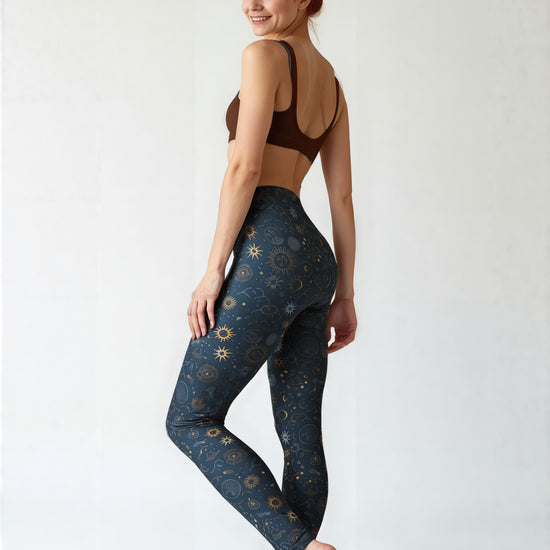 Woman wearing Pixie and Sherbet’s Celestial Luxe Yoga leggings featuring a dark navy base with gold celestial sun, moon, and star print, standing barefoot and smiling in a relaxed pose against a light background.