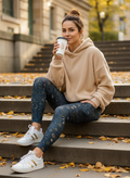 Woman sitting on steps holding a coffee cup, wearing a beige hoodie and blue leggings with white sneakers.