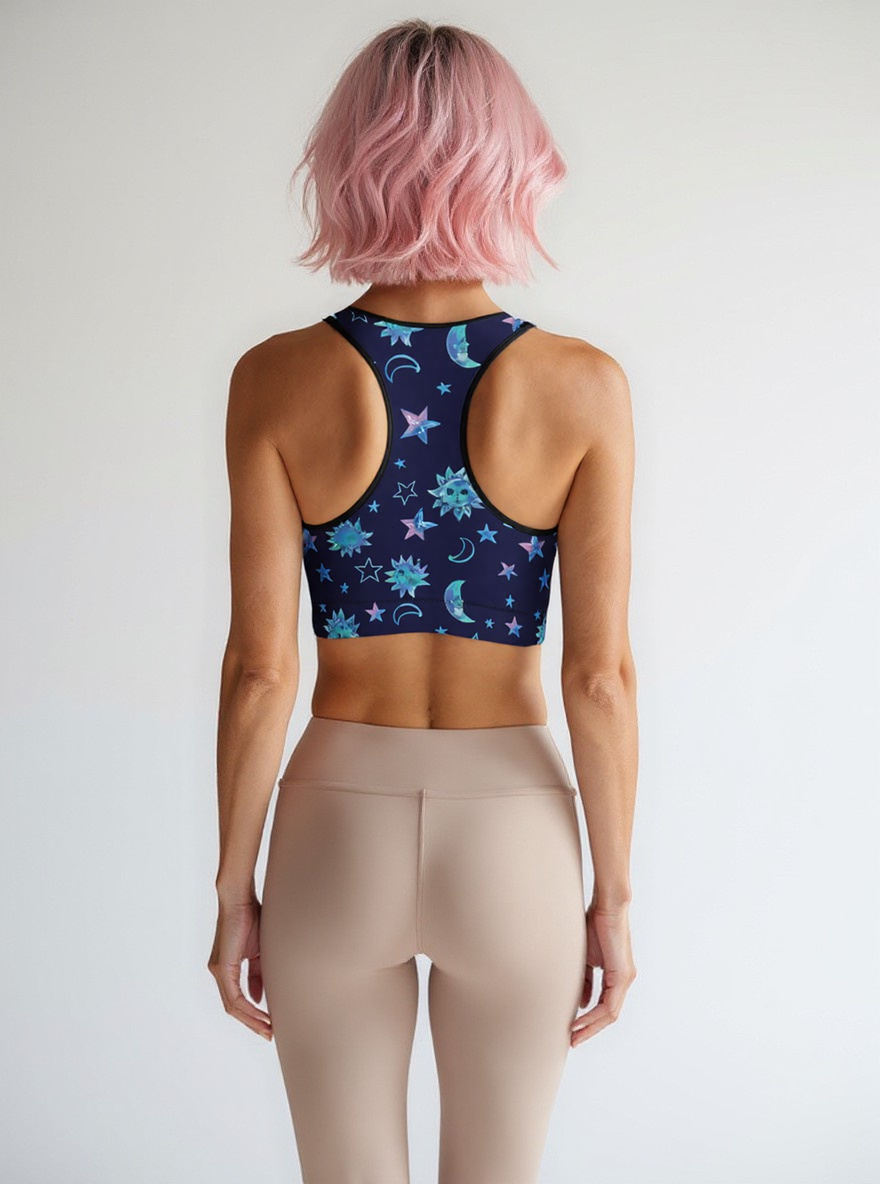 Woman showing the back of the Cosmic Calm Essential Active Bra with a navy base and pastel blue and purple celestial print, standing against a plain background