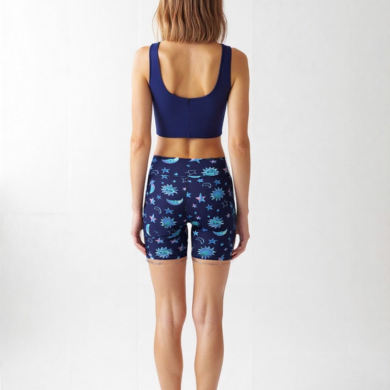 Woman standing barefoot facing away from the camera, wearing a navy blue sports bra and Cosmic Calm yoga shorts with a blue celestial print of stars, moons, and suns, against a plain white background.