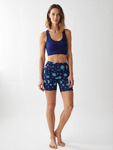 Woman wearing Cosmic Calm yoga shorts featuring a blue celestial pattern with stars, suns, and moons, paired with a solid navy blue sports bra, standing barefoot against a plain white background.