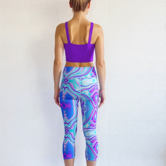 A fair-skinned, athletic woman with blonde hair in a high bun stands barefoot with her back to the camera against a plain white background. She is wearing a bright purple longline sports bra and high-waisted, cropped leggings with a vibrant, swirling blue and purple pattern. Her arms rest naturally at her sides, highlighting the fit and design of the activewear from behind.