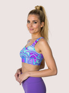 Woman wearing a colourful, swirling pattern "Dreamwave" longline sports bra with purple high-waisted leggings, standing against a plain background and smiling.