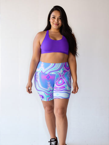Plus-size woman smiling and wearing Dreamwave Yoga Shorts with a vibrant, swirling pattern in blue, purple, and pink, paired with a purple sports bra and black sneakers.
