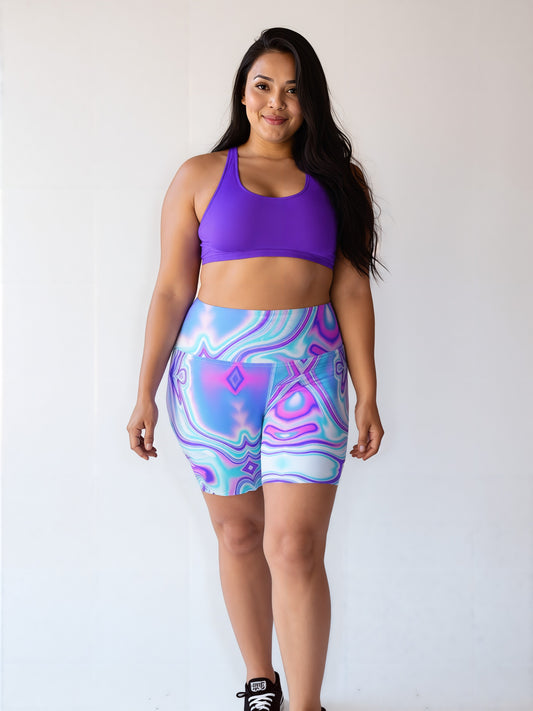 Plus-size woman smiling and wearing Dreamwave Yoga Shorts with a vibrant, swirling pattern in blue, purple, and pink, paired with a purple sports bra and black sneakers.