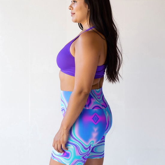 Plus-size woman in profile wearing Dreamwave Yoga Shorts featuring a vibrant blue, purple, and pink swirl pattern, paired with a purple sports bra.