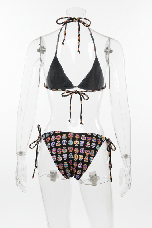 A two-piece string bikini set on a plain white background. The top is solid black with patterned tie straps, and the bottom is black with a colorful sugar skull print.
