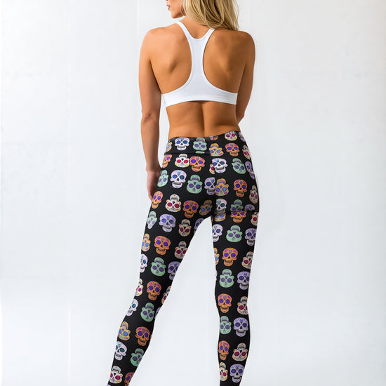 Back view of a woman wearing black leggings with a colourful sugar skull pattern, paired with a white racerback sports bra and black sneakers, standing against a plain background.