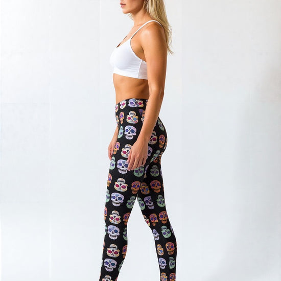 Side view of a woman wearing Eternal Bloom Yoga Leggings with a colourful skull pattern on a black background, paired with a white sports bra and black sneakers, standing against a plain background.