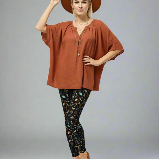 Model wearing Harvest Sky Yoga Leggings with nature-inspired print in yellow, orange, teal, and white on a black base, styled with a rust-coloured V-neck top, brown slip-on shoes, and a wide-brimmed brown hat; outfit blends earthy tones with country-inspired activewear.