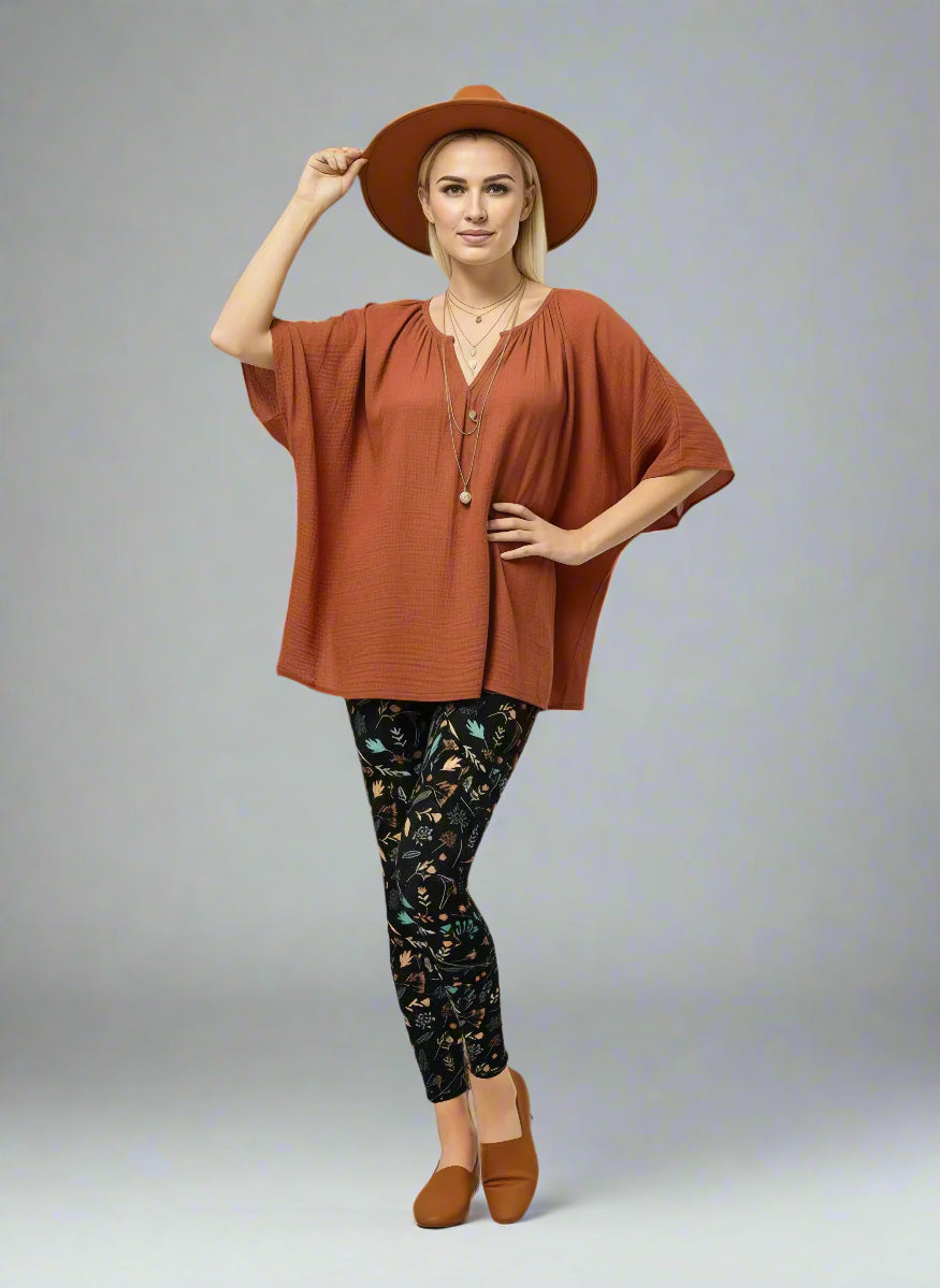 Model wearing Harvest Sky Yoga Leggings with nature-inspired print in yellow, orange, teal, and white on a black base, styled with a rust-coloured V-neck top, brown slip-on shoes, and a wide-brimmed brown hat; outfit blends earthy tones with country-inspired activewear.