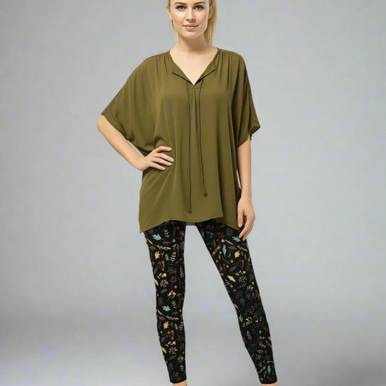 Model wearing Harvest Sky Yoga Leggings with black base and nature-inspired print in orange, teal, yellow, and white, styled with an olive green short-sleeve blouse with tie detail and light brown slip-on shoes; casual everyday look blending earthy tones with country-inspired activewear.