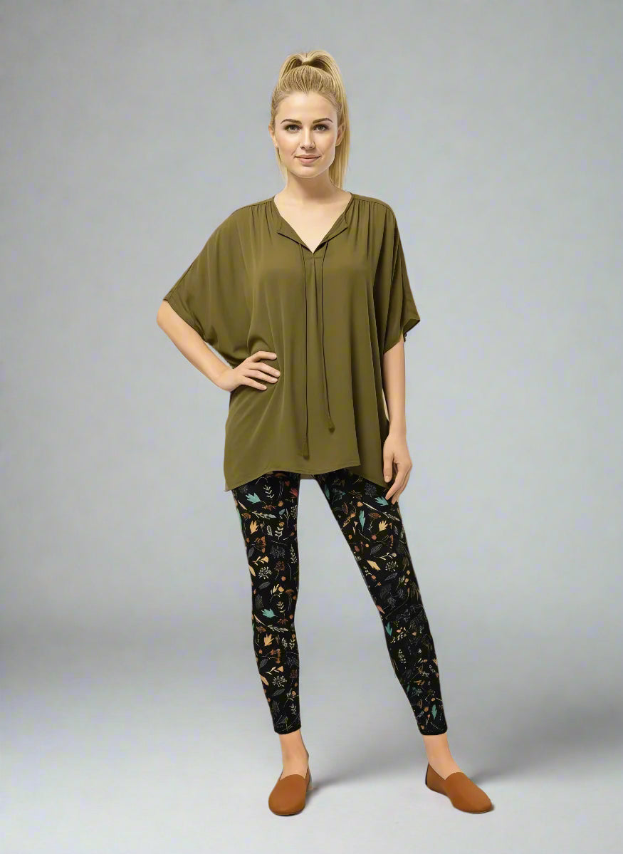 Model wearing Harvest Sky Yoga Leggings with black base and nature-inspired print in orange, teal, yellow, and white, styled with an olive green short-sleeve blouse with tie detail and light brown slip-on shoes; casual everyday look blending earthy tones with country-inspired activewear.