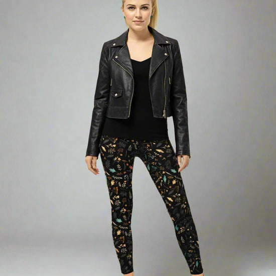 Model wearing Harvest Sky Yoga Leggings with black base and nature-inspired print in orange, teal, yellow, and white, styled with a black leather jacket, fitted black top, and brown slip-on shoes; outfit blends edgy streetwear with country-inspired activewear.