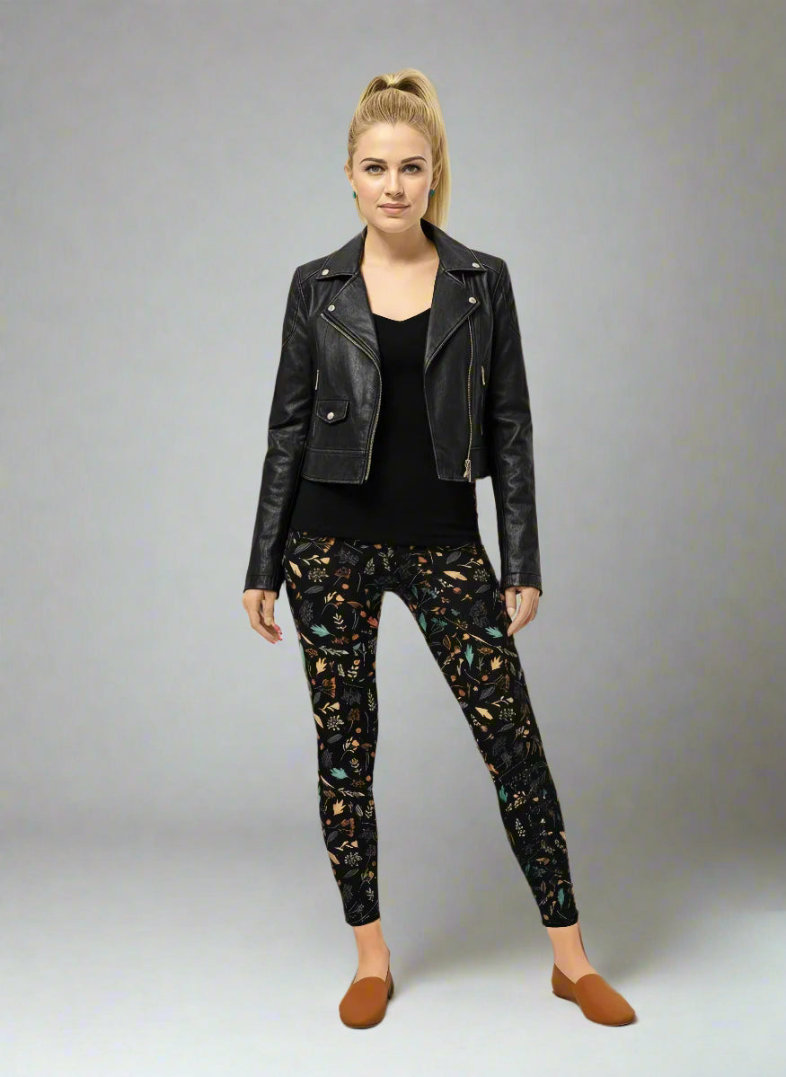 Model wearing Harvest Sky Yoga Leggings with black base and nature-inspired print in orange, teal, yellow, and white, styled with a black leather jacket, fitted black top, and brown slip-on shoes; outfit blends edgy streetwear with country-inspired activewear.