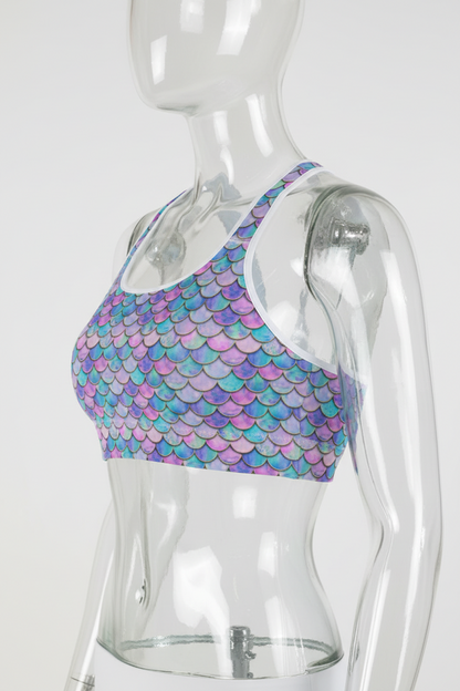 A woman wearing a sports bra with a colorful mermaid scale pattern in shades of blue, purple, and pink, paired with white leggings, shown against a plain white background.