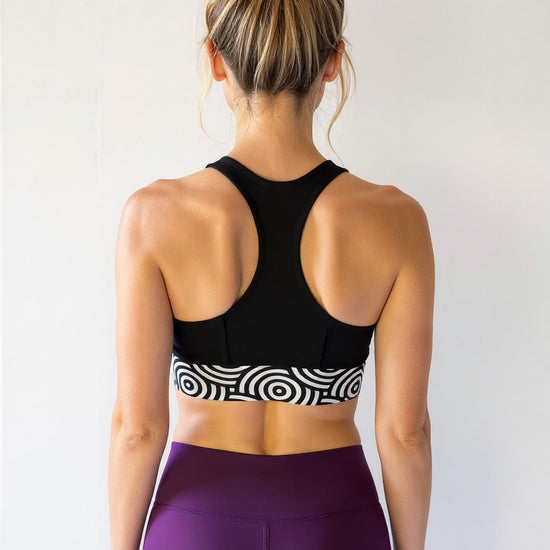 Athletic woman with blonde hair in a messy bun, shown from the back. She is wearing a black longline sports bra with a bold black and white circular "Lunar Loops" print band and high-waisted deep purple leggings, standing against a plain light background.